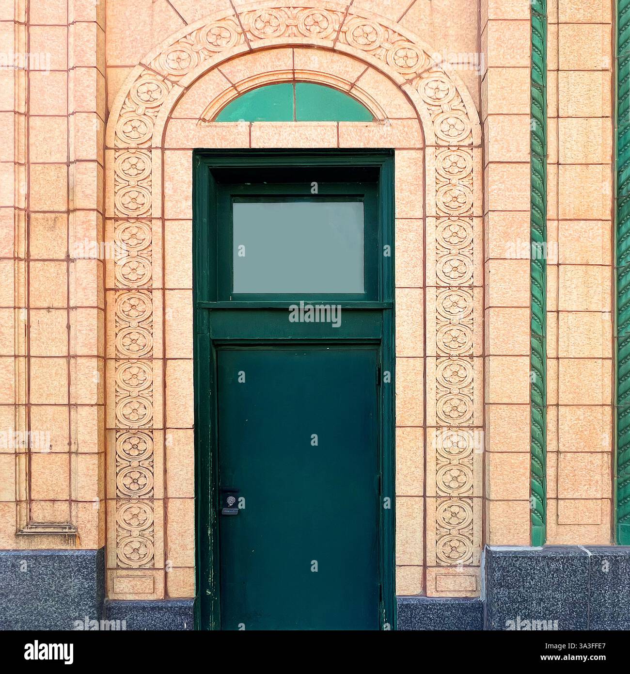 Eine kunstvoll verzierte grüne Tür innerhalb einer wunderschön detaillierten Terrakotta-Fassade mit komplizierten Blumenschnitzereien und einem bogenförmigen Heckfenster. - Smartphone-aufgenommenes Stockfoto