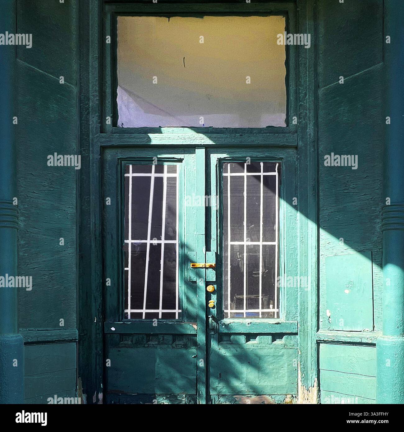 Alte grüne Ladentür mit Gitterglaspaneelen, teilweise von Sonnenlicht beleuchtet, zeigt eine verwitterte Fassade in einem urbanen Ambiente. Stockfoto Alte grüne Ladentür mit Gitterglaspaneelen, teilweise von Sonnenlicht beleuchtet, zeigt eine verwitterte Fassade in einem urbanen Ambiente. Stockfoto
