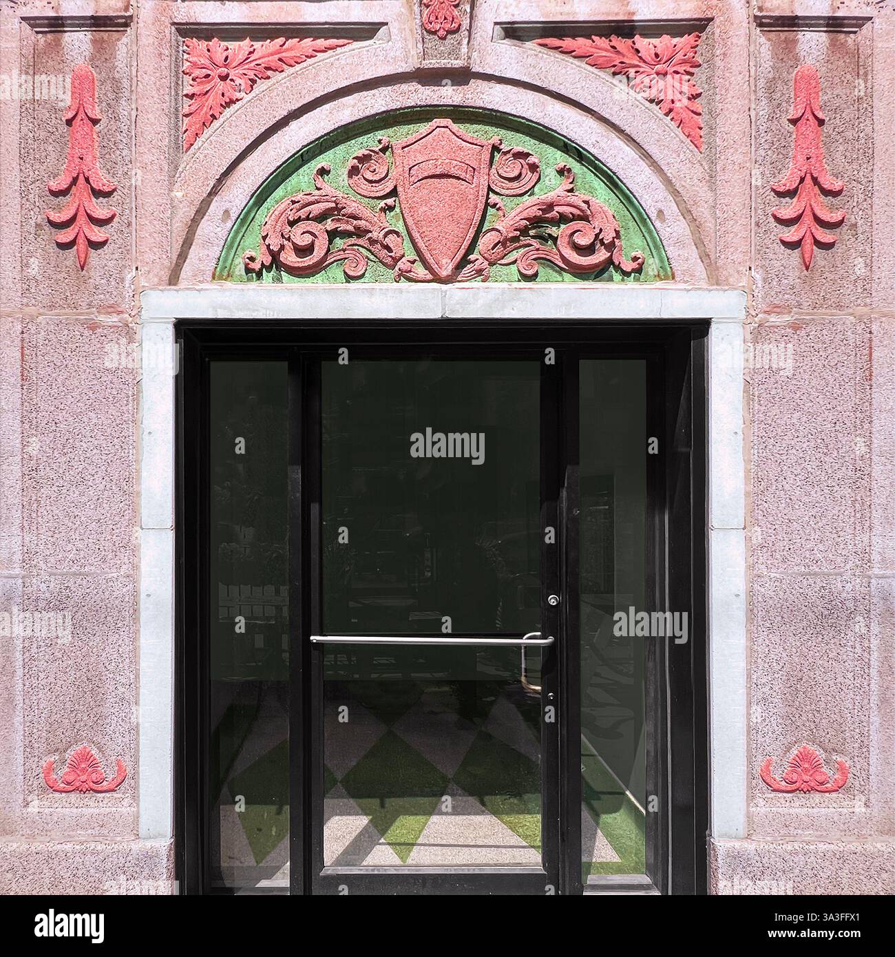 Eleganter Eingang mit einem kunstvollen Steinbogen mit verzierten roten und grünen dekorativen Details, die eine moderne Glastür innerhalb einer historischen Fassade einrahmen. Stockfoto Eleganter Eingang mit einem kunstvollen Steinbogen mit verzierten roten und grünen dekorativen Details, die eine moderne Glastür innerhalb einer historischen Fassade einrahmen. Stockfoto