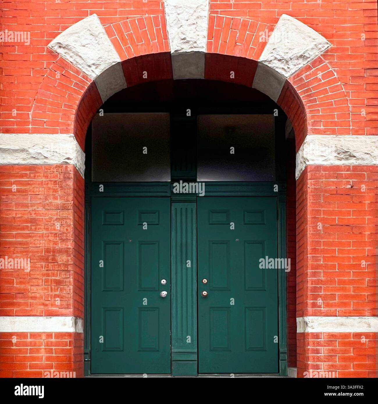 Bogenförmiger Eingang mit tiefgrünen Doppeltüren in einer roten Ziegelfassade mit auffälligen Kalksteindetails. Stockfoto
