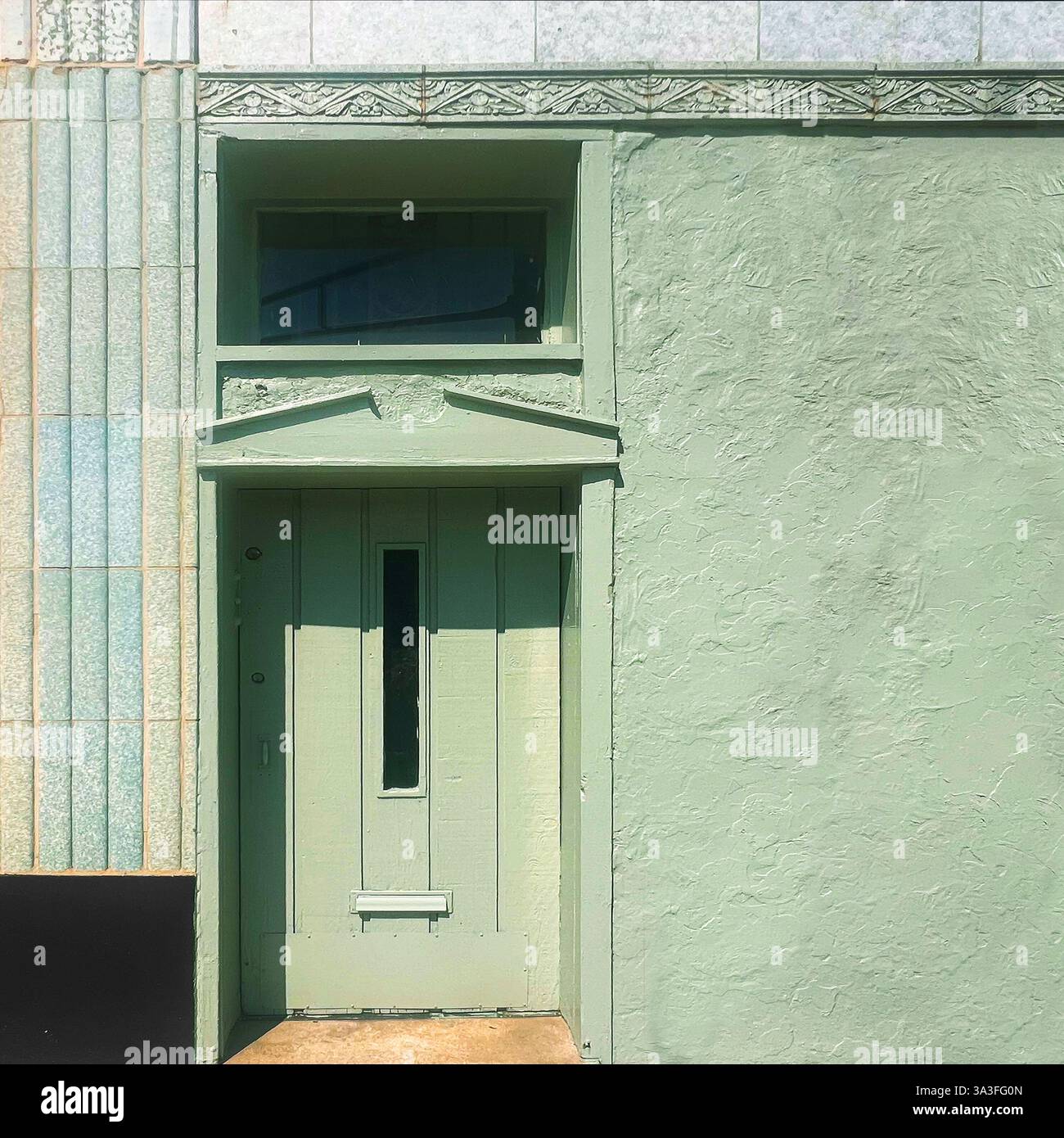Eine pastellgrüne Holztür mit einem schmalen vertikalen Fenster, eingerahmt von einer strukturierten Stuckfassade und dekorativen architektonischen Details, die in Sonnenlicht getaucht sind. Stockfoto