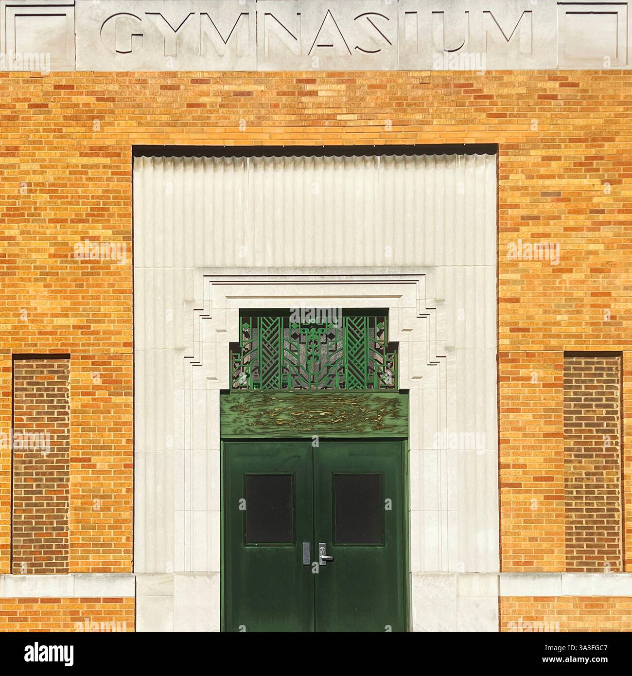 Eingang des Art déco-Sporthauses mit dunkelgrünen Metalltüren, dekorativem geometrischem Kühlergrill und detailliertem Mauerwerk vor einer klassischen Ziegelfassade. - Smartphone-aufgenommenes Stockfoto