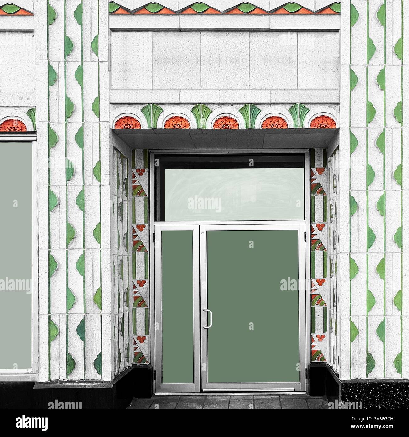 Art déco-Eingang mit polierten Metall- und Glastüren, die von verzierten grünen und orangen geometrischen und floralen Motiven auf weißem Stein eingerahmt werden. Stockfoto Art déco-Eingang mit polierten Metall- und Glastüren, die von verzierten grünen und orangen geometrischen und floralen Motiven auf weißem Stein eingerahmt werden. Stockfoto