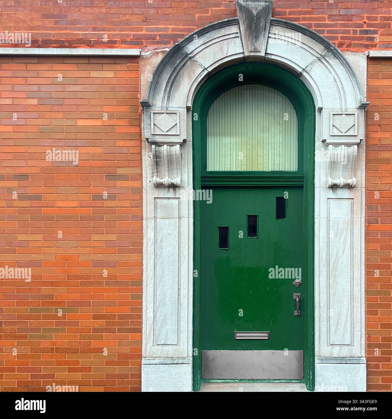 Tür mit einer bogenförmigen grünen Holztür mit einzigartigen asymmetrischen Fenstern, eingerahmt von einer lebendigen orangen Ziegelfassade mit Steinschnitzereien. - Smartphone-aufgenommenes Stockfoto