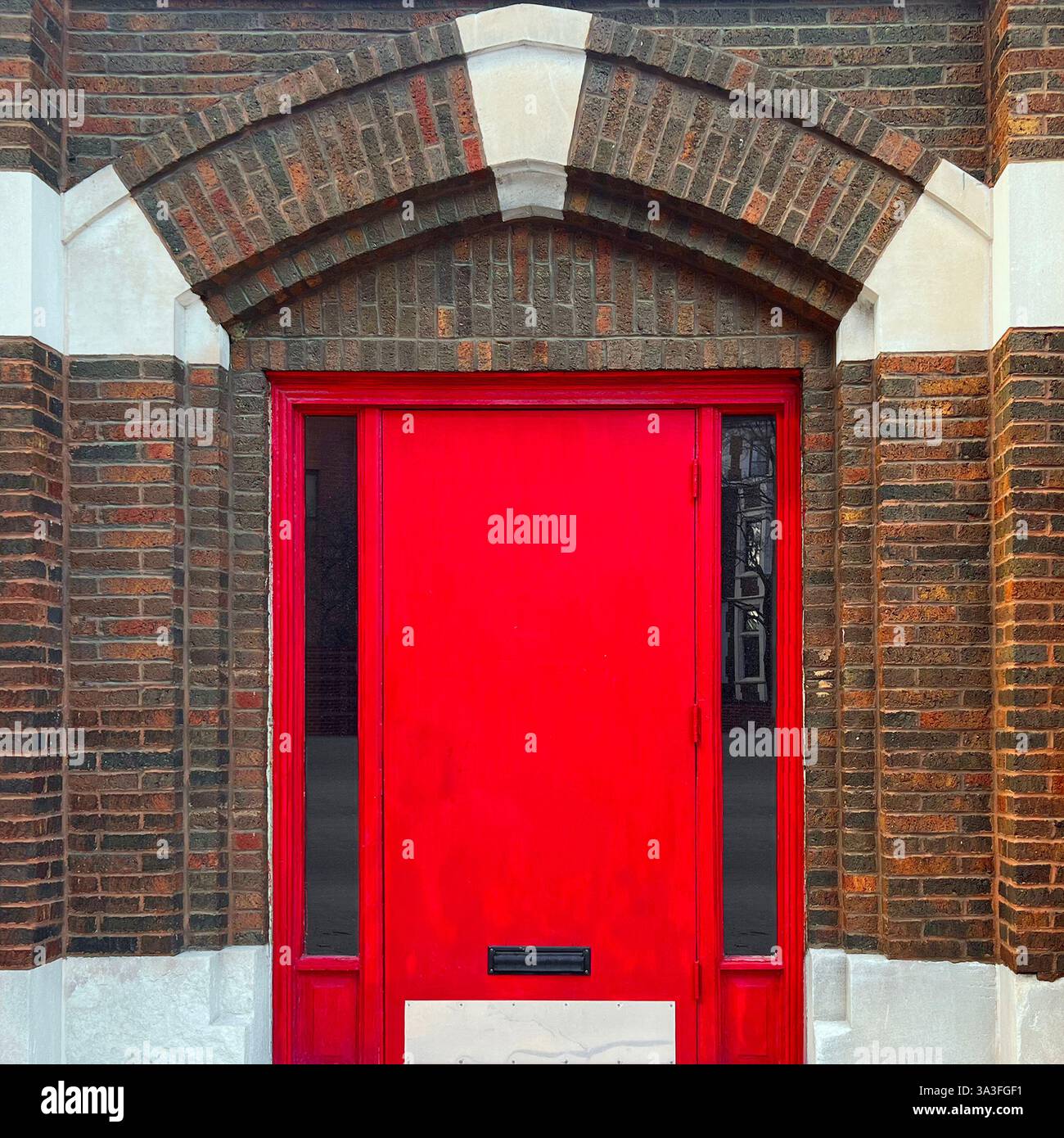 Rote Tür vor einer braunen Ziegelfassade, eingerahmt von einem dramatischen gotischen Bogen mit kontrastierenden Steinakzenten. - Smartphone-aufgenommenes Stockfoto