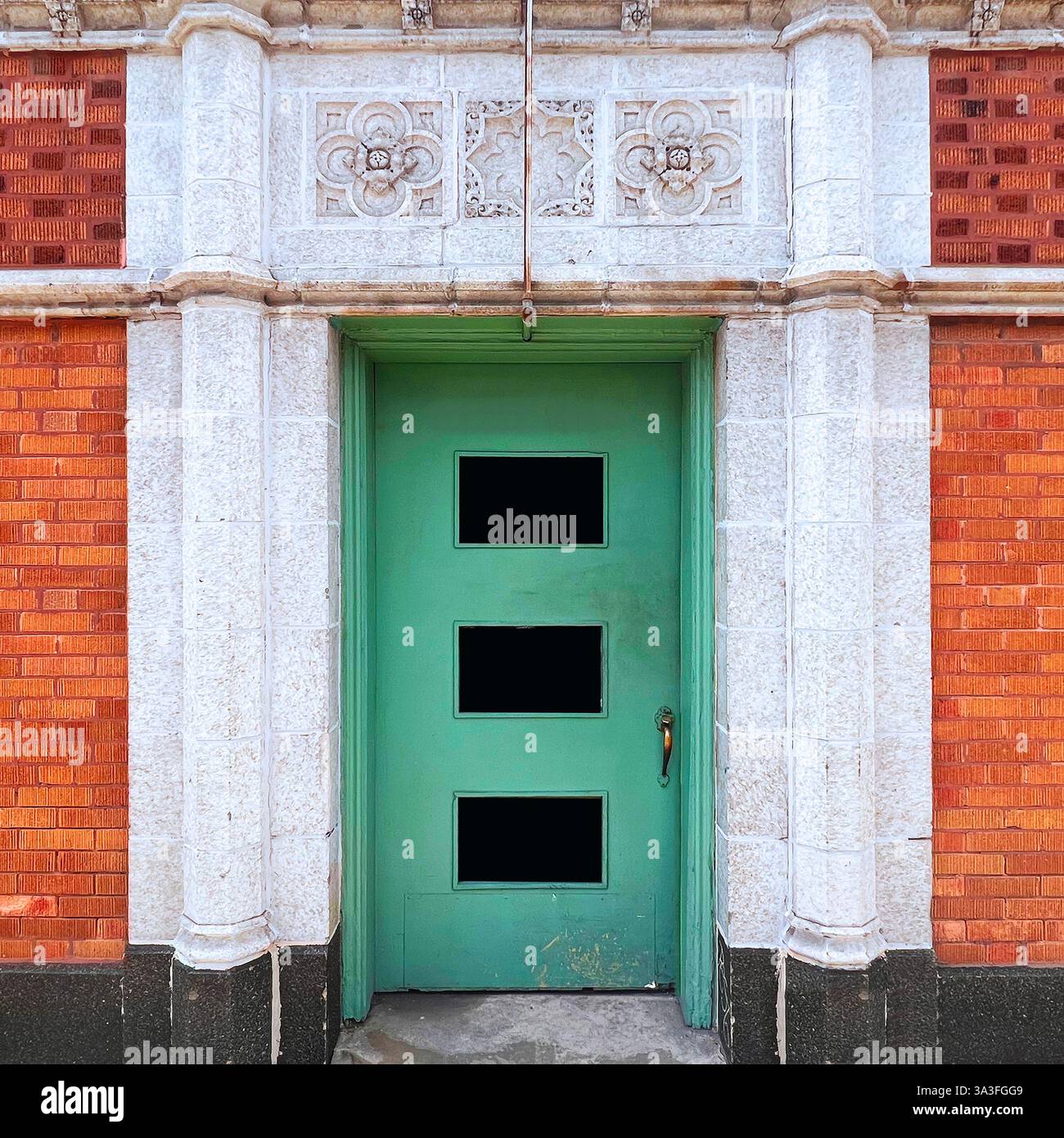 Mintgrüne Metalltür mit drei ausgeschnittenen Fenstern, die in einer kunstvollen Fassade aus weißem Stein und orangen Ziegelsteinen mit komplizierten Blumenschnitzereien eingefasst sind. - Smartphone-aufgenommenes Stockfoto