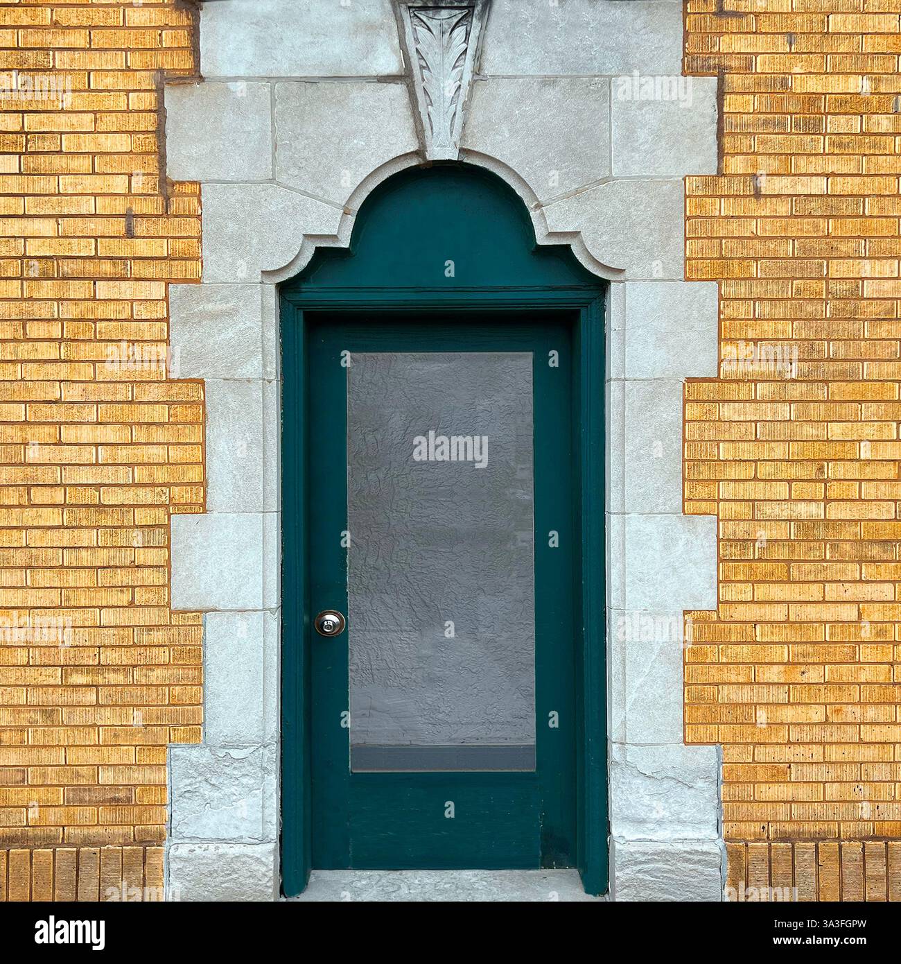 Dunkelgrüne Tür mit Milchglaspaneel in einem kunstvollen Steinbogen, eingerahmt von einer gelben Ziegelfassade mit dekorativen Steindetails, Chicago - Smartphone-aufgenommenes Stockfoto