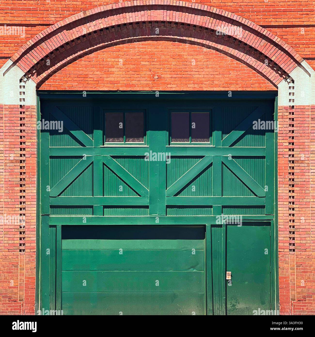 Eine große grüne Vintage-Lagertür mit klassischem X-Brace-Design in einem historischen roten Backsteingebäude mit einem Bogengemauerwerk. Stockfoto