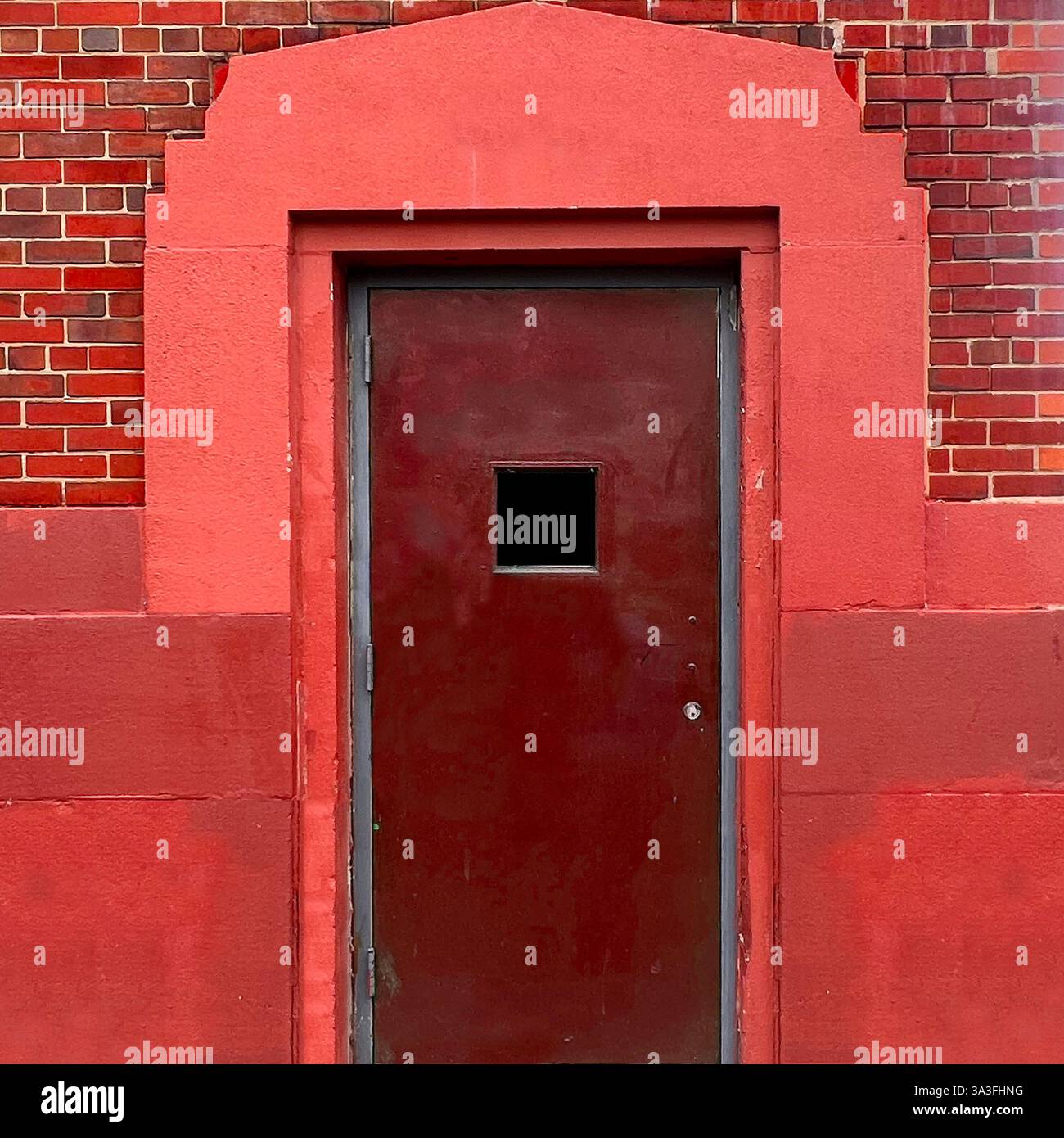 Eine dunkelrote Stahltür mit einem kleinen quadratischen Fenster steht in einer Chicagoer Gasse von kräftigem rotem Mauerwerk und reichem Mauerwerk eingerahmt. - Smartphone-aufgenommenes Stockfoto
