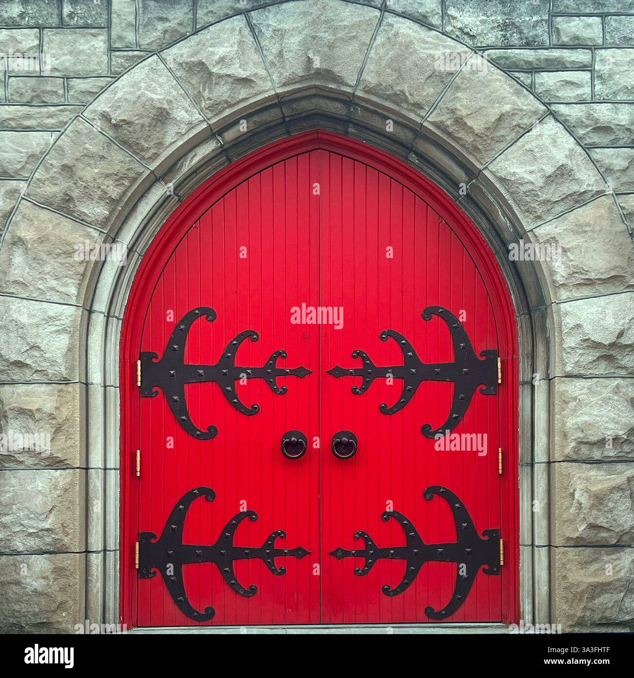 Rote Kirchtüren im gotischen Stil mit verzierten schwarzen schmiedeeisernen Scharnieren, die in einem großen Steinbogen angeordnet sind. - Smartphone-aufgenommenes Stockfoto