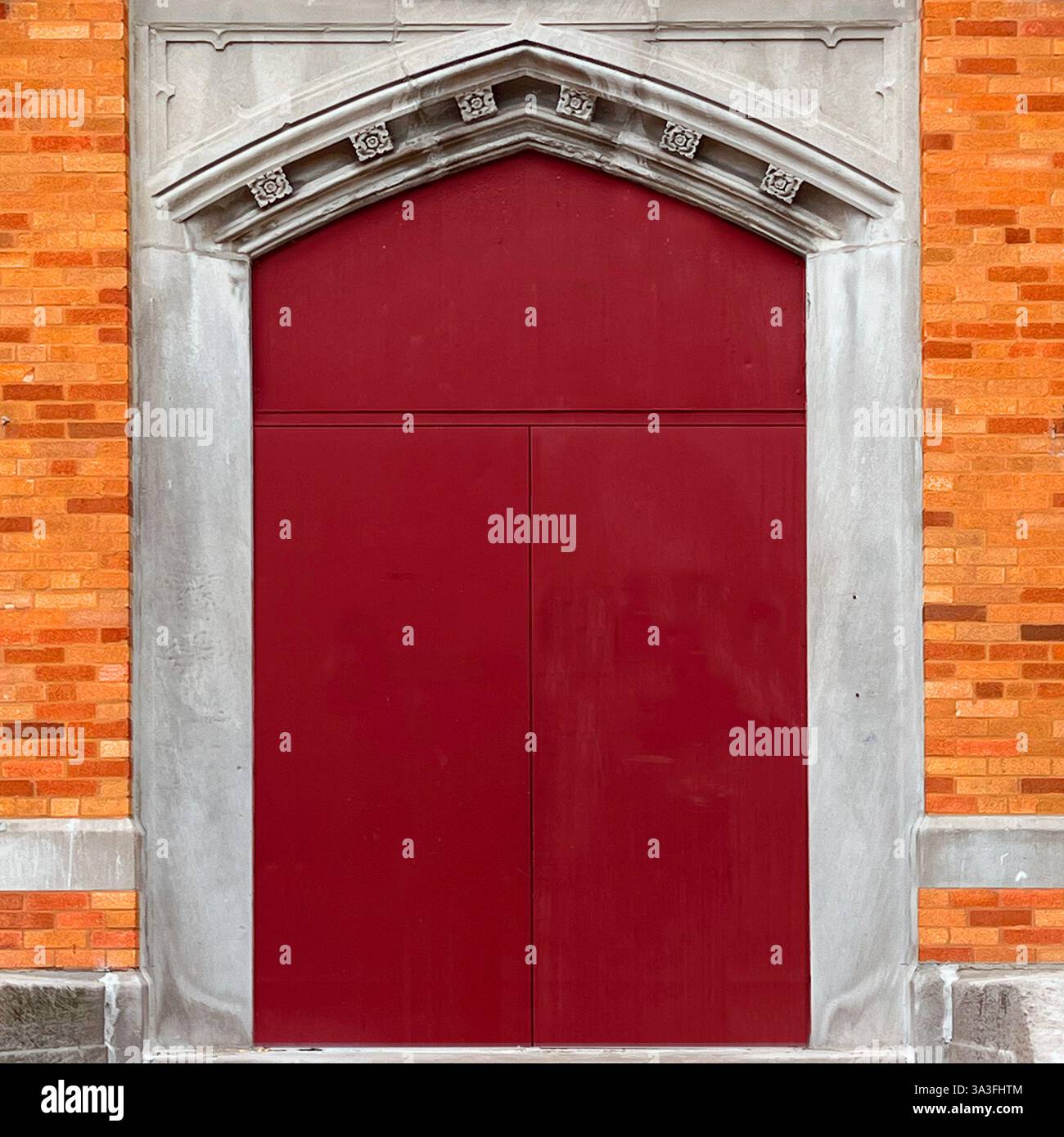 Rote Doppeltür in einem gotischen Steinbogen vor einer warmen Ziegelfassade - Smartphone-aufgenommenes Stockfoto