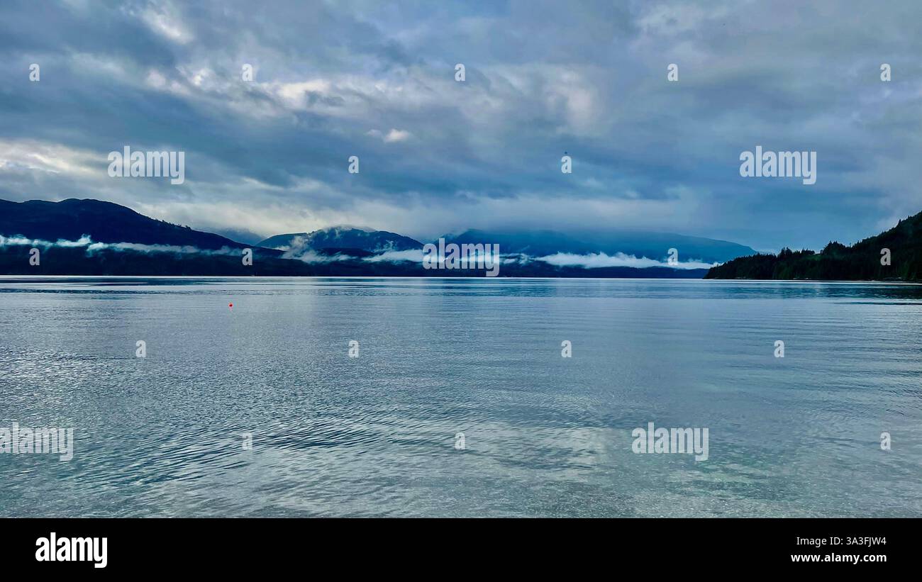 kanadas Flagge am Ufer eines Flusses mit einem trockenen Stock und grünen Bergen im Hintergrund - Smartphone-aufgenommenes Stockfoto