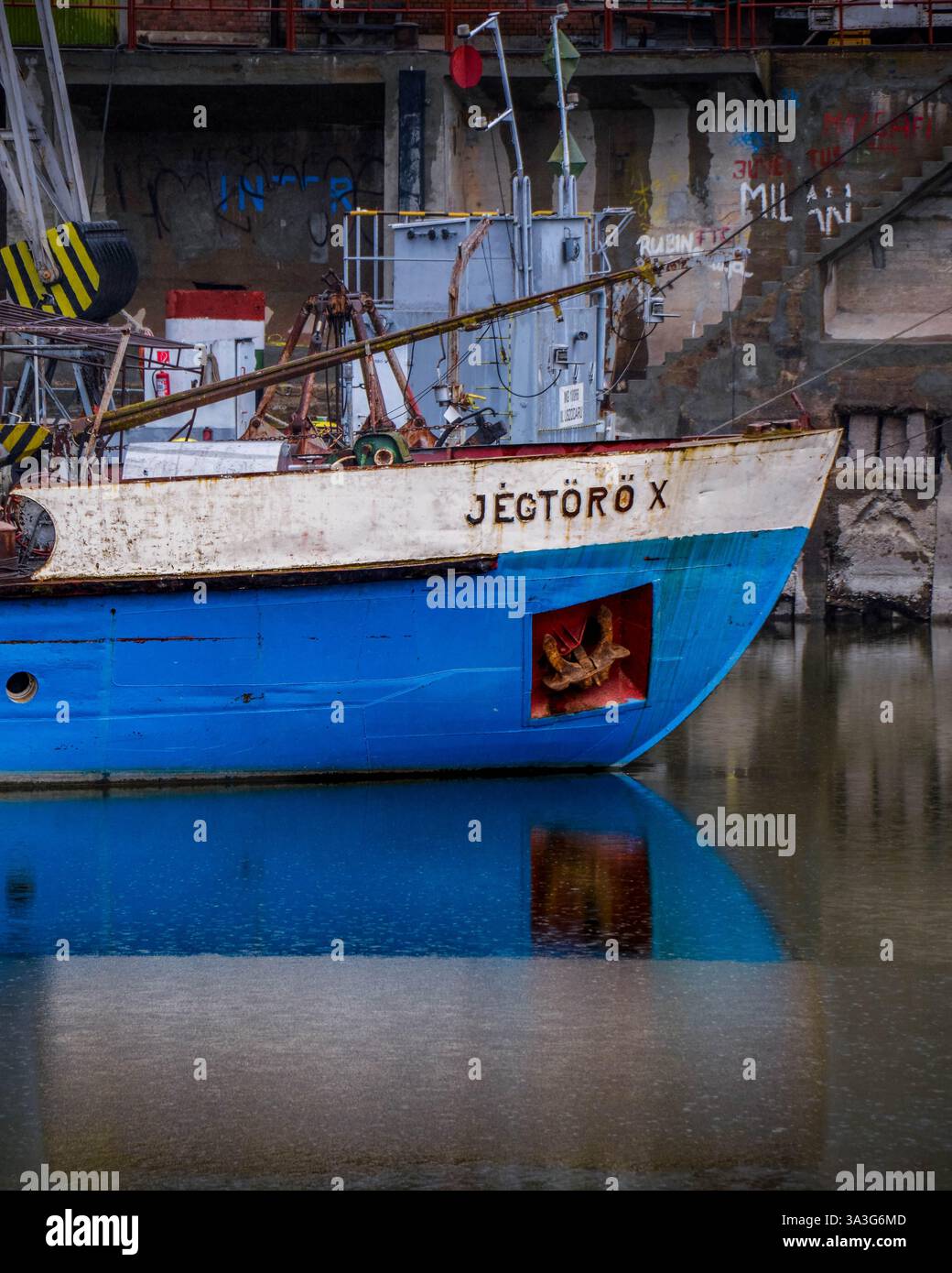 Das blau-weiße Eisbrecherschiff „JÉGTÖRŐ X“ legte an einem Hafen an, dessen rostiger Rumpf sich auf ruhigen Gewässern vor einer industriellen Kulisse spiegelt. Stockfoto