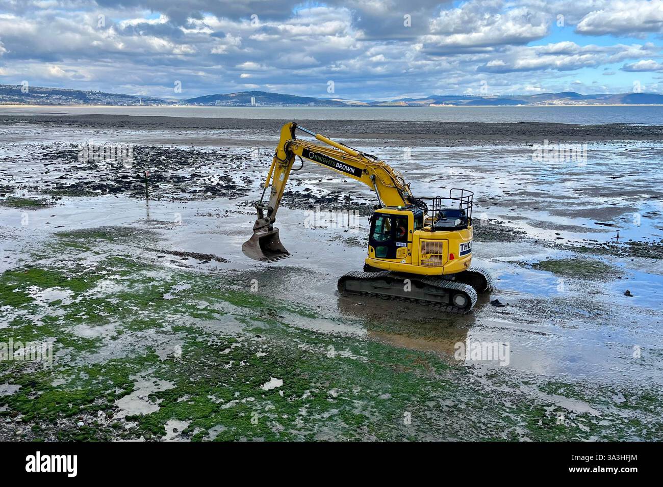 Gelber Bagger, der am Strand in Mumbles entlang fährt, mit Swansea Bay im Hintergrund. Murbles, Swansea, Wales, Vereinigtes Königreich. März 2025. Stockfoto Gelber Bagger, der am Strand in Mumbles entlang fährt, mit Swansea Bay im Hintergrund. Murbles, Swansea, Wales, Vereinigtes Königreich. März 2025. Stockfoto