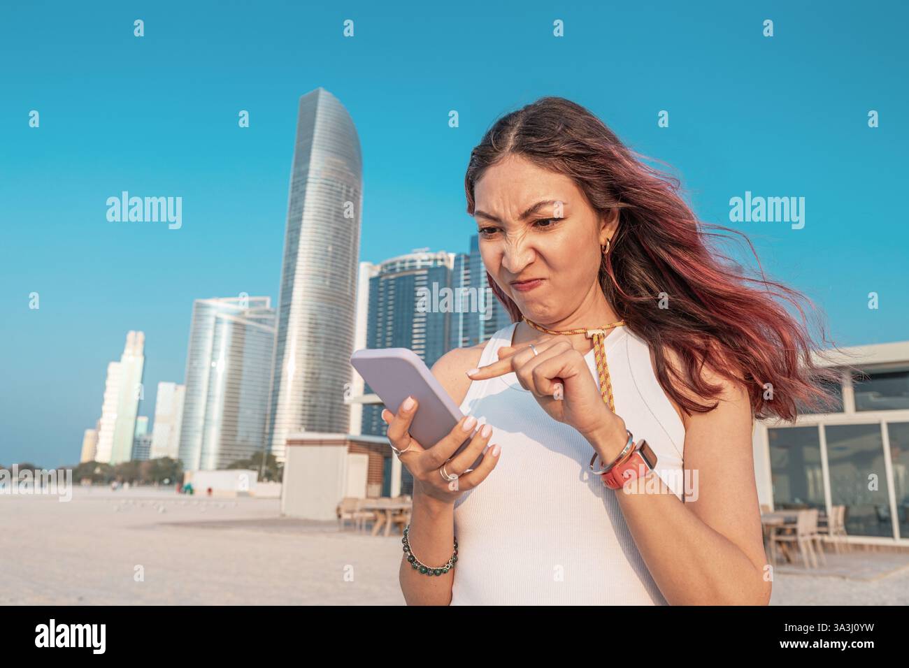 Eine junge Frau, die die Stirn runzelte, während sie Nachrichten auf ihrem Smartphone liest und vor den Wolkenkratzern in der sonnigen Stadt Abu Dhabi steht Stockfoto