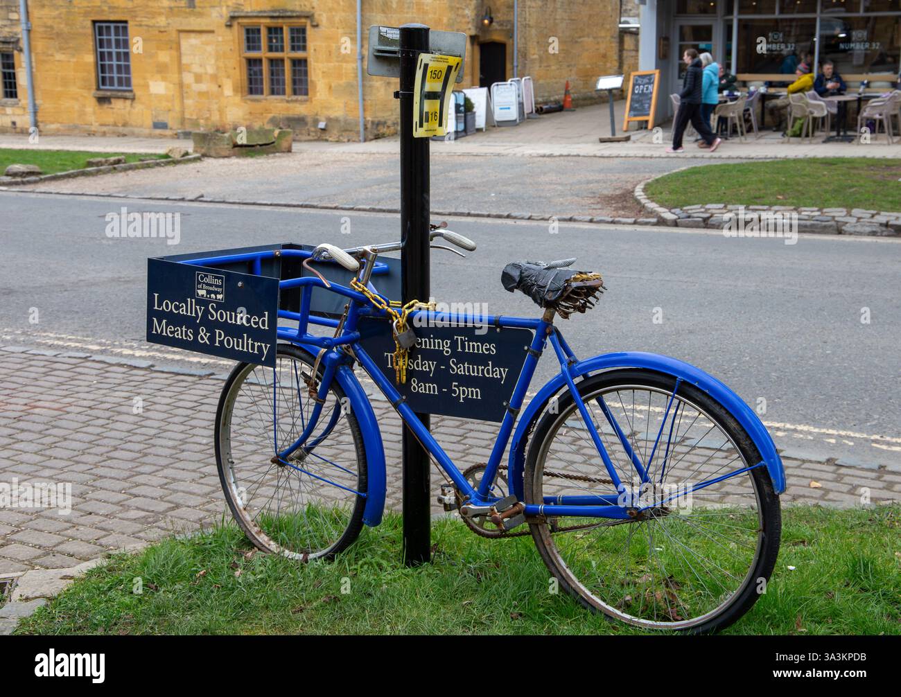 Blaues Vintage-Fahrrad mit einem Lieferkorb, der neben einem Schild geparkt wird, auf dem lokale Fleischwaren & Geflügel Broadway Cotswold's UK gewarnt wird Stockfoto