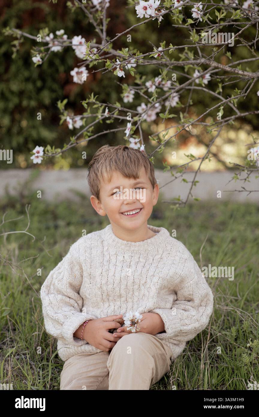 Ein Junge sitzt unter einem blühenden Mandelbaum, pflückt frische Mandeln und hält sie in seinen Händen. Stockfoto