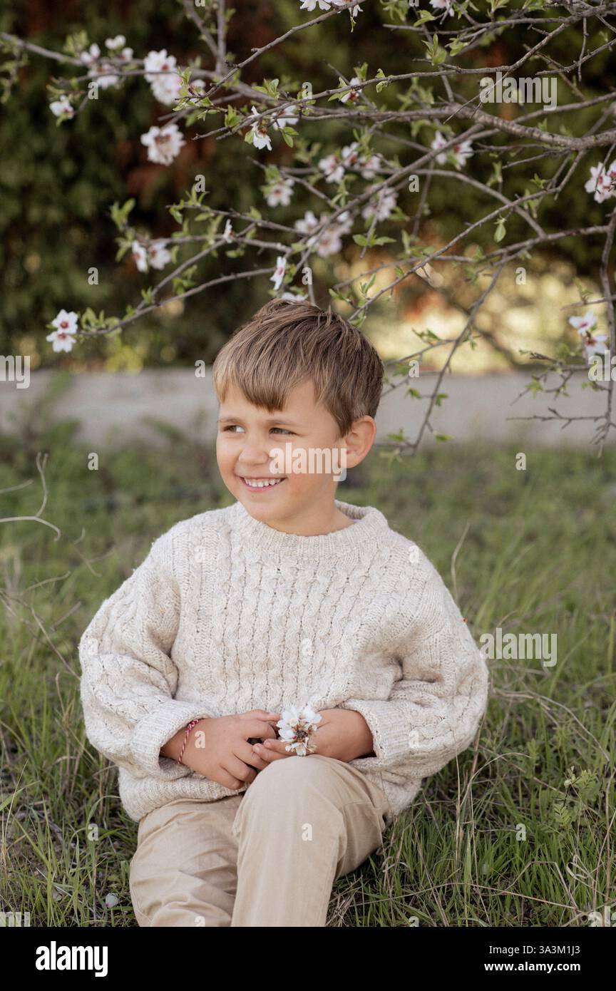Ein Junge sitzt unter einem blühenden Mandelbaum, pflückt frische Mandeln und hält sie in seinen Händen. Stockfoto