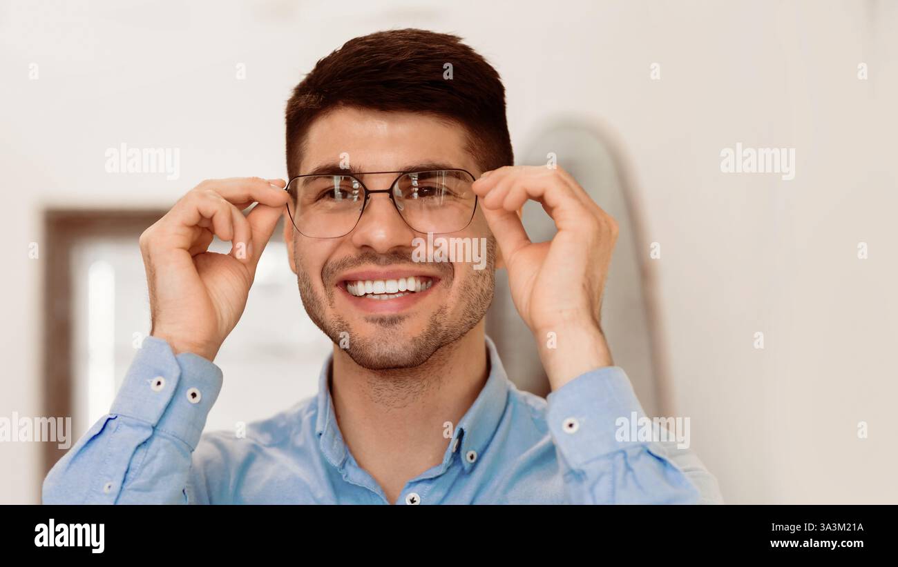 Ein junger Mann passt seine Brille an und lächelt in einem hellen Innenraum Stockfoto