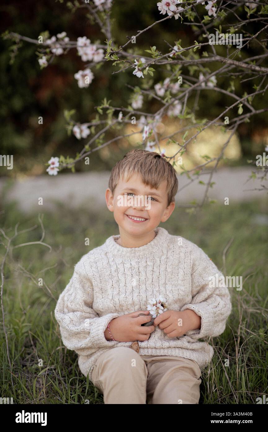 Ein Junge sitzt unter einem blühenden Mandelbaum, pflückt frische Mandeln und hält sie in seinen Händen. Stockfoto