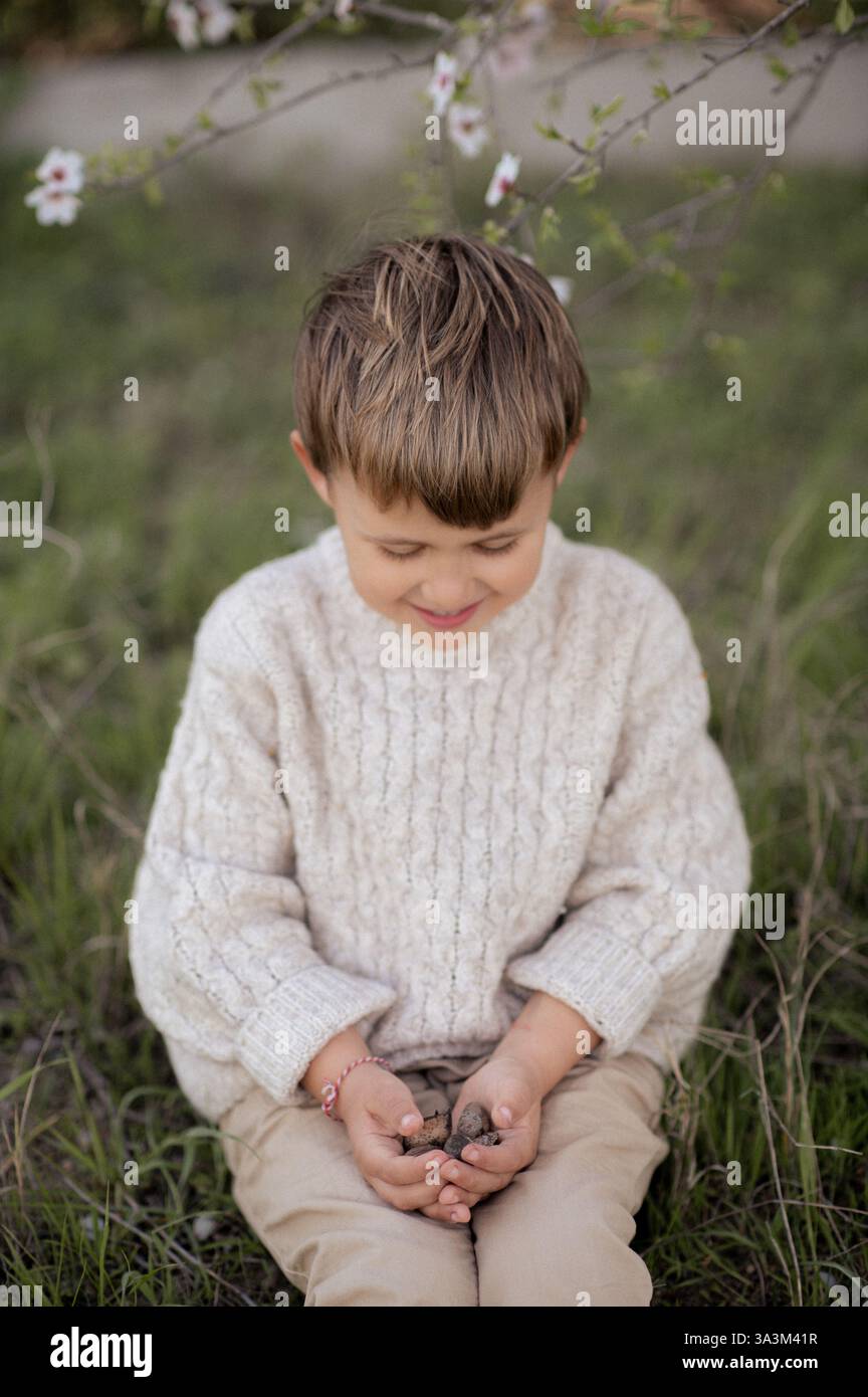 Ein Junge sitzt unter einem blühenden Mandelbaum, pflückt frische Mandeln und hält sie in seinen Händen. Stockfoto