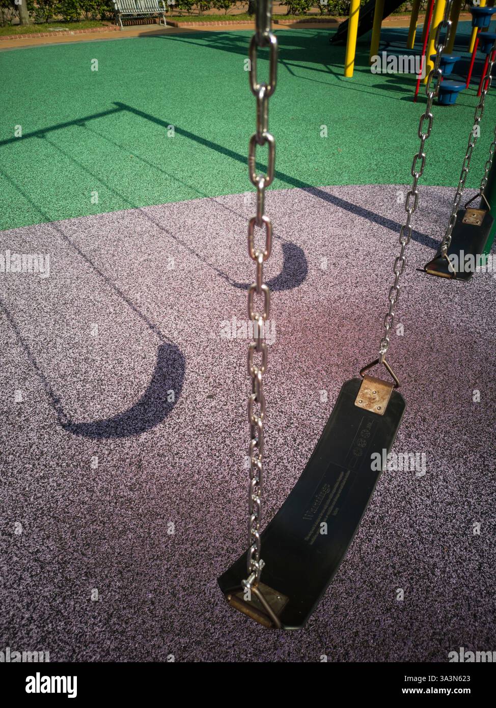 Ein sonniger Tag wirft Schatten auf die Schaukel. Gepolsterter Bodenbelag aus recycelten Materialien mit strapazierfähiger spezieller Gummibeschichtung für mehr Sicherheit Stockfoto
