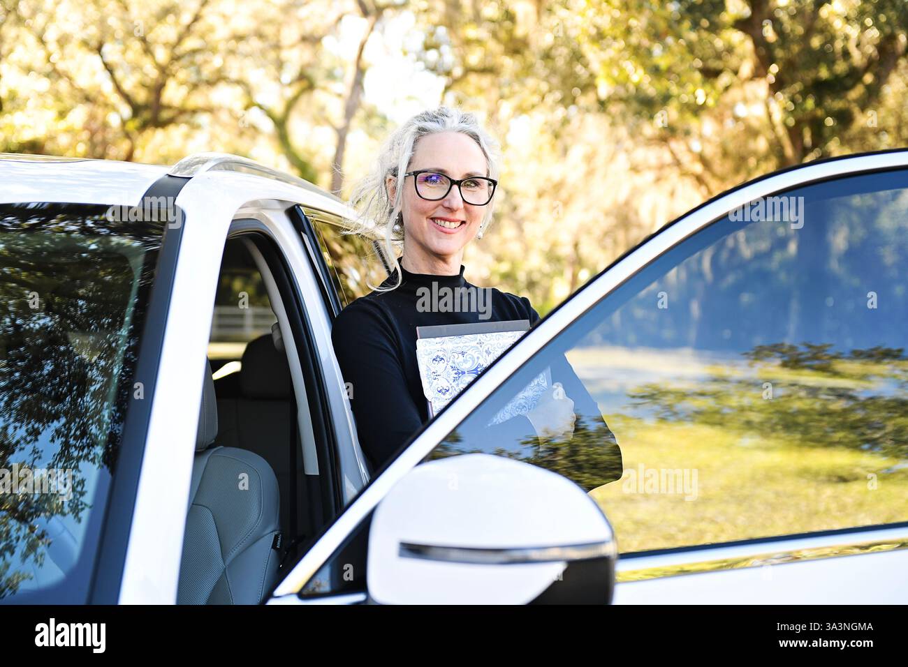 Frau mit Brille, die sich auf einen weißen SUV stützt, Bücher hält, Outdoo Stockfoto