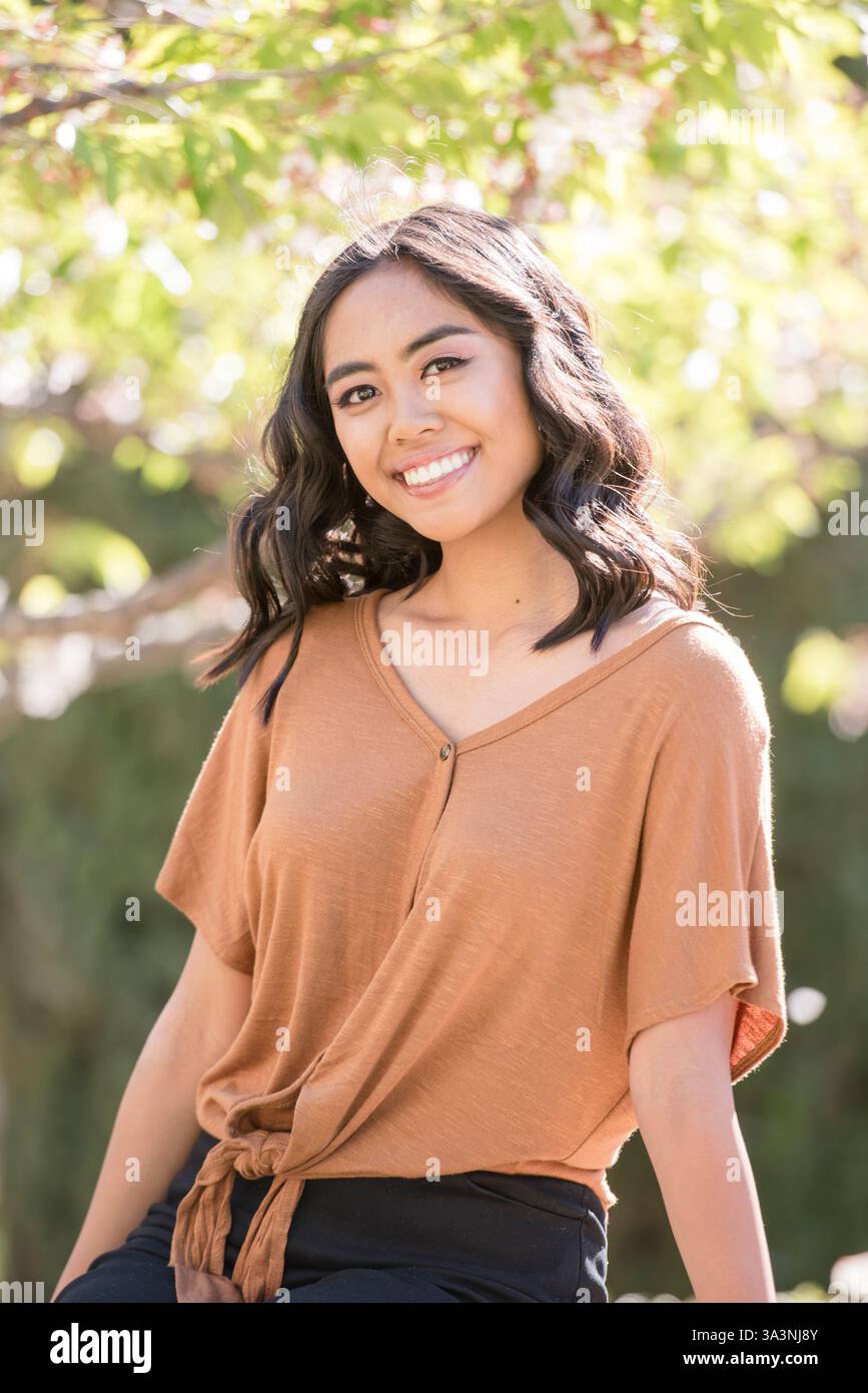 Die junge Frau lächelt warm, während sie draußen unter sonnendurchfluteten Bäumen sitzt Stockfoto