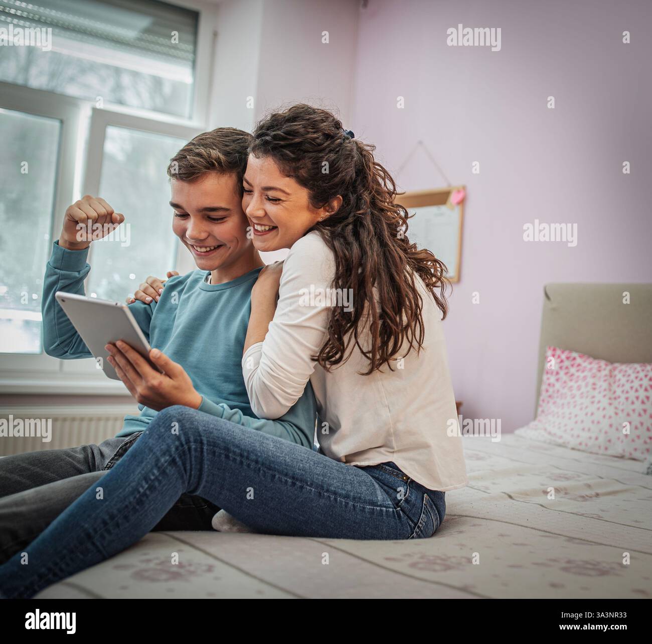 Mutter und Sohn teilen aufregende Momente, während sie Inhalte auf einem Tablet erkunden und bequem auf dem Bett in einem gemütlichen, in sanften Farben dekorierten Schlafzimmer sitzen Stockfoto