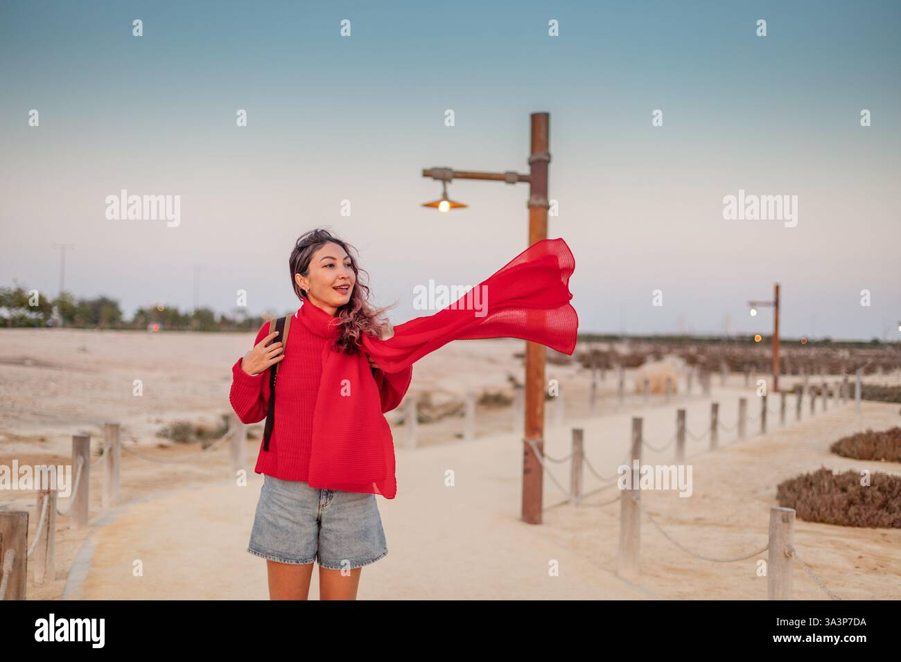 Eine junge Frau in roter Kleidung, die ihren Schal hält, wird bei Sonnenuntergang vom Wind auf einem Fußweg am Strand geblasen Stockfoto