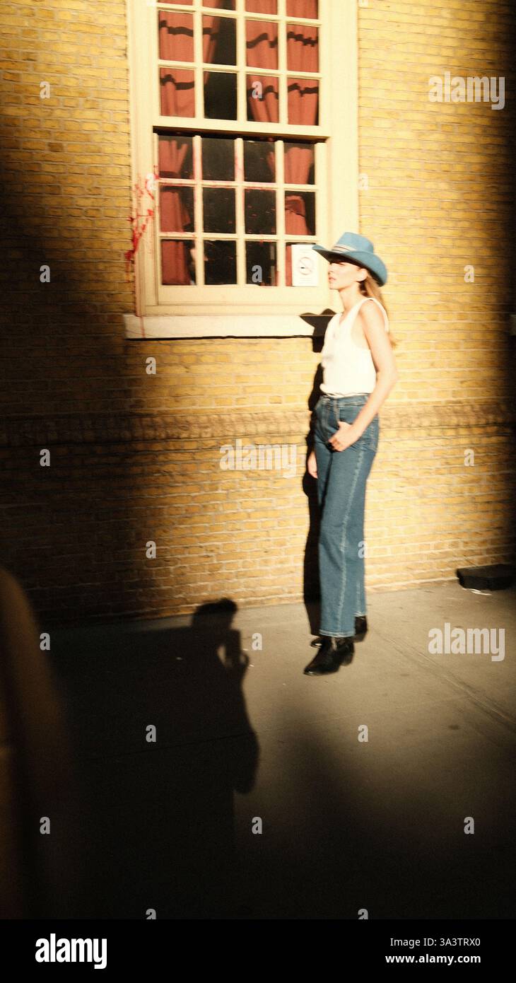 Eine Frau mit blauem Cowboyhut steht vor einer sonnendurchfluteten Ziegelwand und wirft einen scharfen Schatten. Das goldene Stundenlicht hebt die warmen Töne hervor, während das Rot- Stockfoto