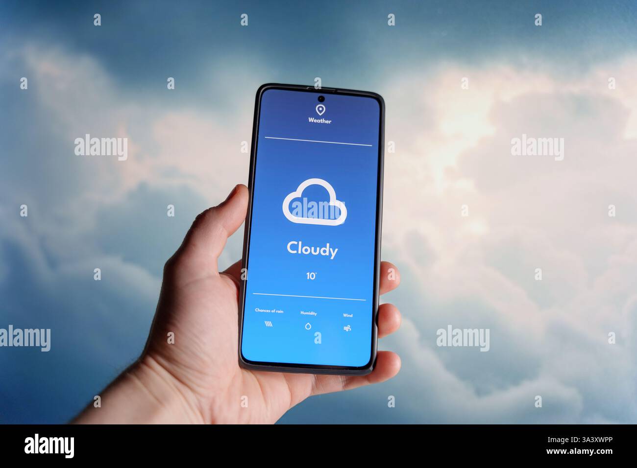 Hand hält ein Smartphone, das eine Wetter-App mit einer bewölkten Vorhersage anzeigt. Weiche Wolken füllen den Himmel im unscharfen Hintergrund. Wettertechnologie A Stockfoto