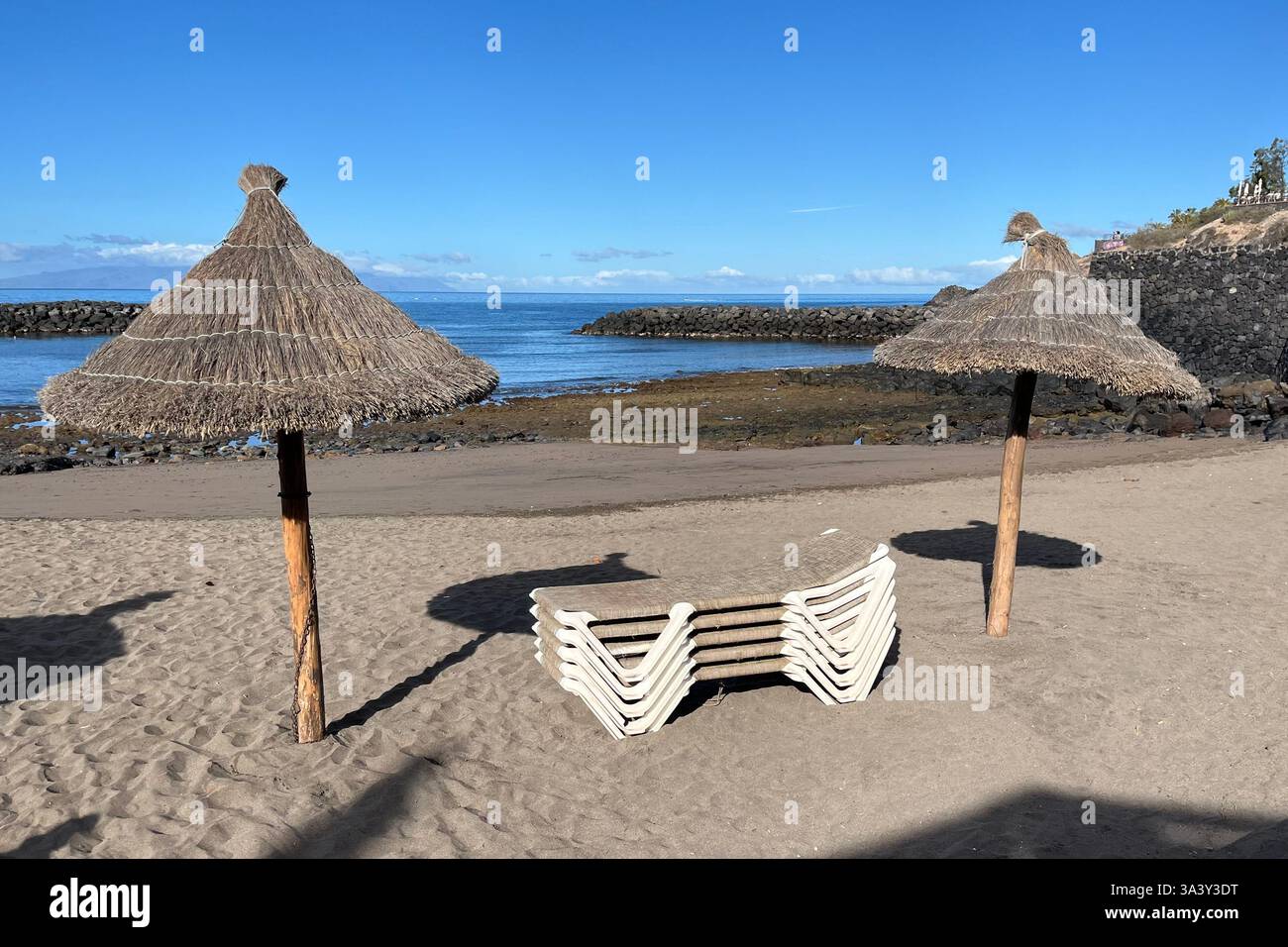 Ein Stapel Liegestühle zwischen zwei Strohschirmen am Strand El Bobo. Costa Adeje, Teneriffa, Kanarische Inseln, Spanien. Februar 2025. - Smartphone-aufgenommenes Stockfoto