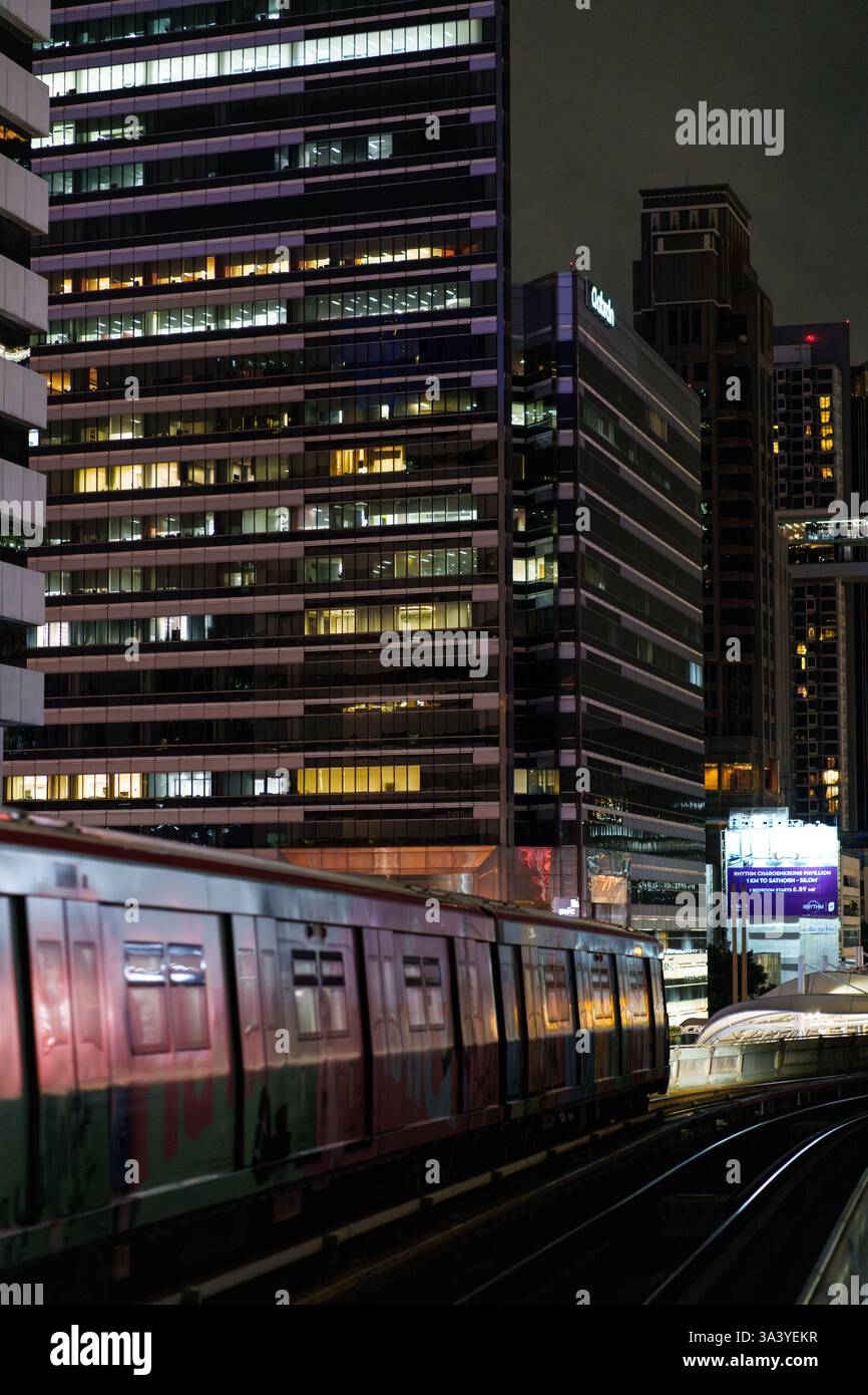 Dieses nächtliche Bild zeigt einen BTS Skytrain, der durch Bangkok, Thailands Geschäftsviertel, fährt, umgeben von beleuchteten Wolkenkratzern. Das Glühen Stockfoto