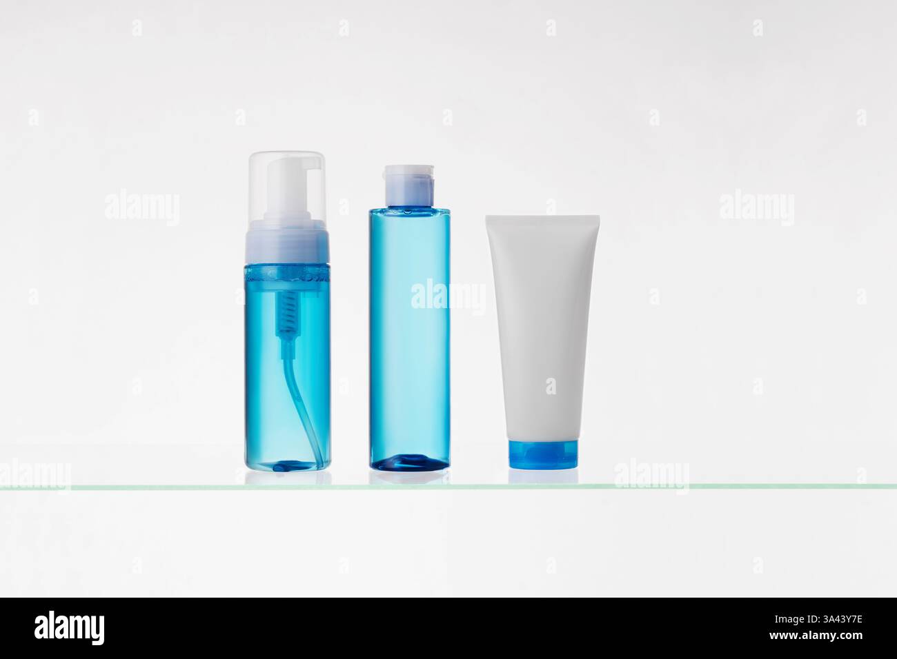 Blaue transparente Flaschen und weiße Röhre mit kosmetischen Produkten auf einem Glasregal auf weißem Hintergrund. Mockup von Hautpflegeprodukten Stockfoto
