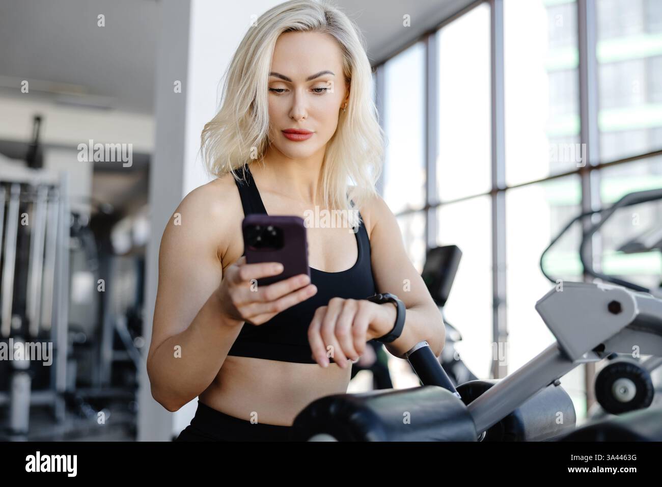 Blonde Athletin, die Fitness-App auf dem Smartphone nutzt und Smartwatch während des Workouts im Fitnessstudio überprüft Stockfoto