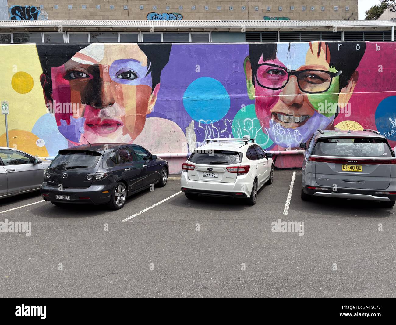 Porträtgemälde auf einer Gebäudewand hinter geparkten Autos auf einem Parkplatz, mit einem farbenfrohen und abstrakten Design. Stockfoto
