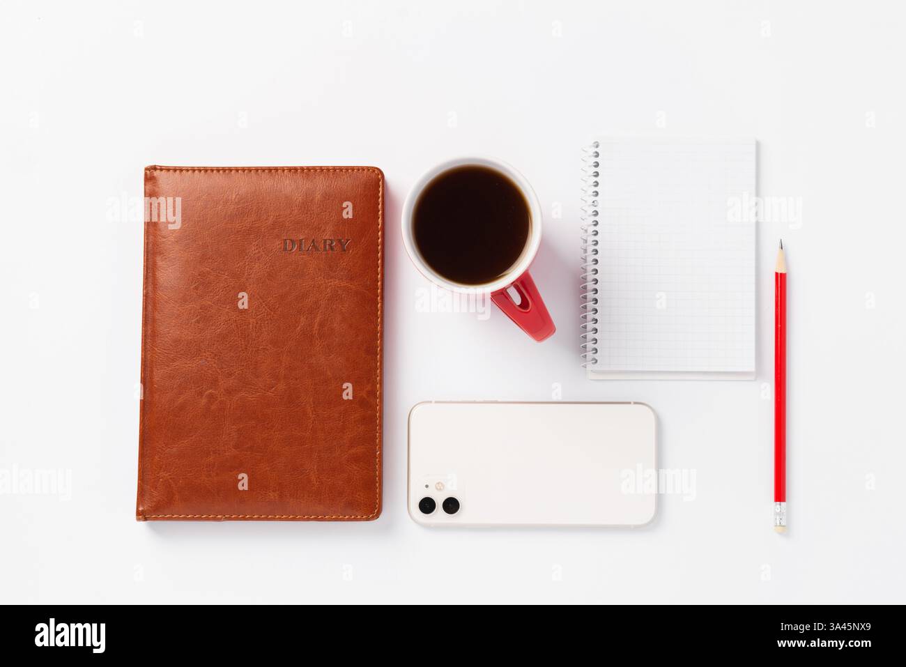 Braunes Notizbuch, roter Bleistift, weißes Smartphone und rote Tasse mit Kaffee auf weißem Hintergrund. Morgendämmerpläne für den Tag, Arbeitsplatz Stockfoto