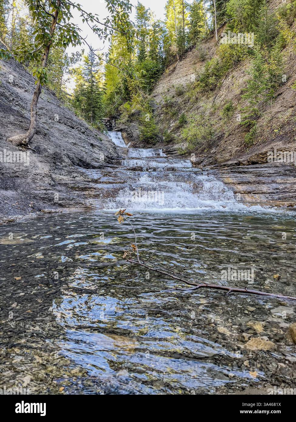 Ein kleiner Wasserfall fließt über felsiges Gelände, umgeben von grünen Bäumen und zerklüfteten Klippen. Das klare Wasser an der Basis reflektiert das f - Smartphone-aufgenommenes Stockfoto