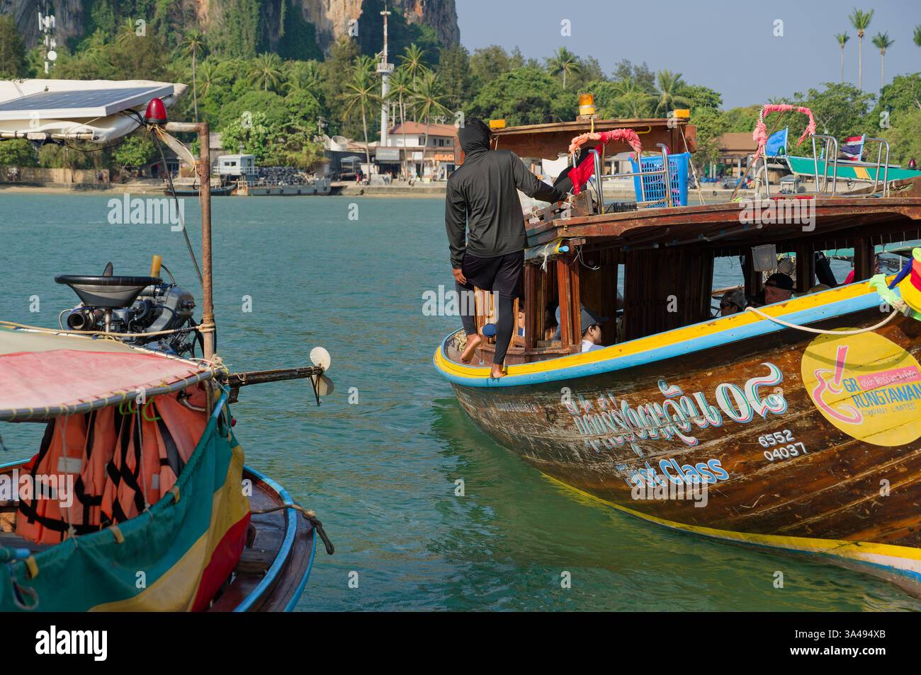 Ein maskierter Bootsarbeiter hält sich barfuß auf einem hölzernen Langboot am schwimmenden Pier der East Railay Stockfoto