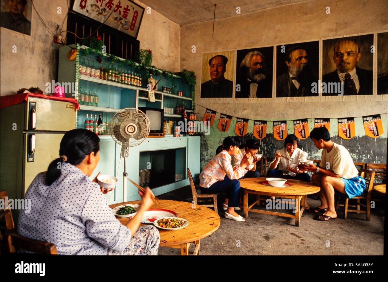 Menschen essen in einem Restaurant im ländlichen China in der Nähe von Huanghuacheng mit Plakaten von Mao, Lenin und anderen marxistischen Führern und verkaufen Batterien. Stockfoto