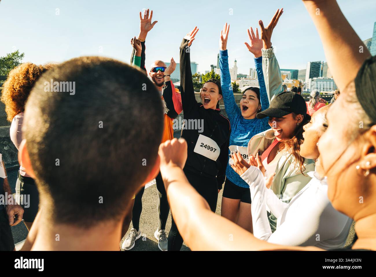 Eine vielfältige Gruppe begeisterter Läufer feiert nach einem Halbmarathon. Die Umgebung ist städtisch, die Teilnehmer heben ihre Hände, zeigen Stockfoto
