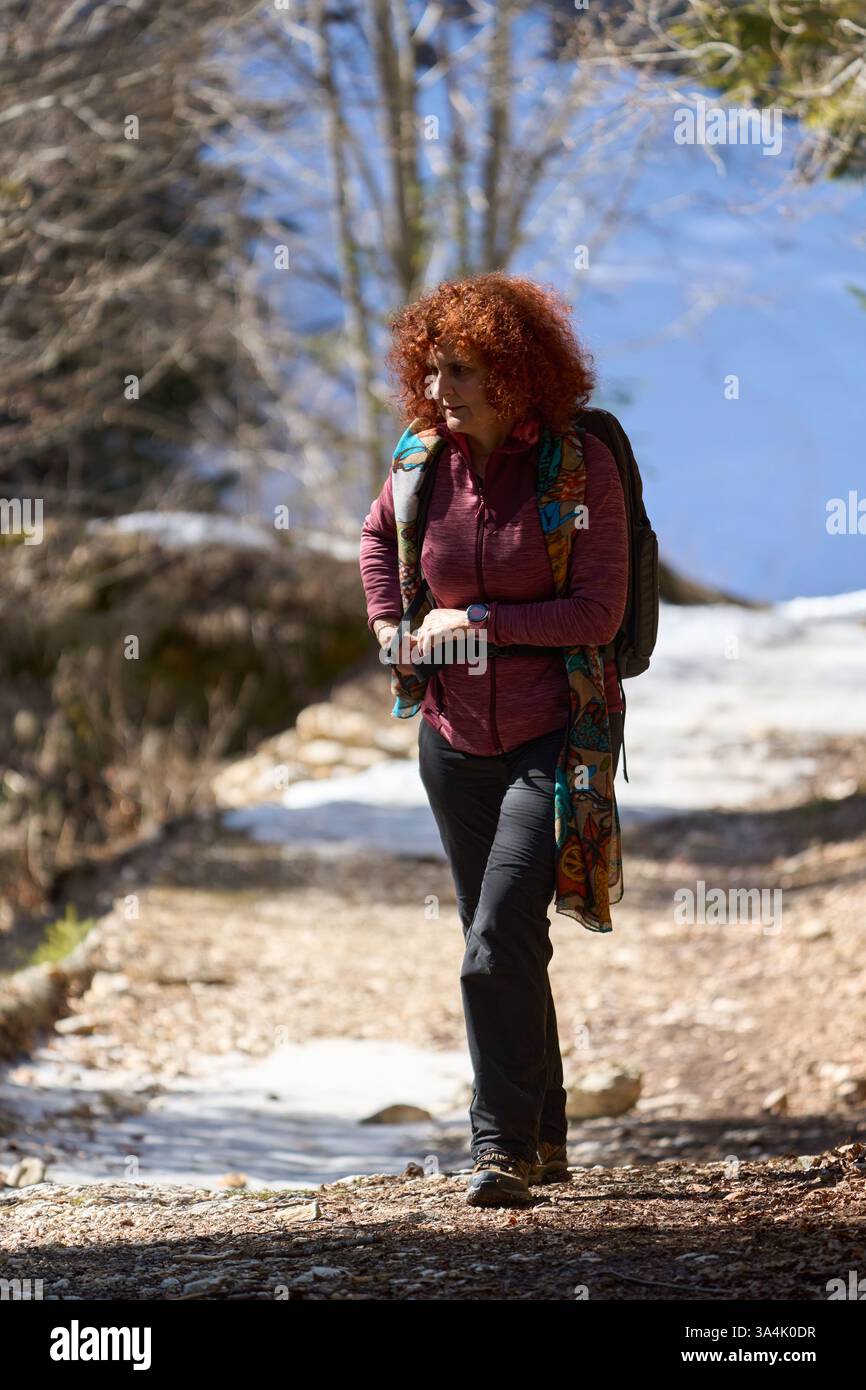Frau mit lockigen roten Haaren wandert auf einem Waldweg mit Schneeflächen in hellem Sonnenlicht Stockfoto