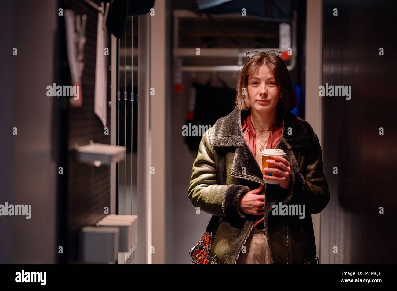 Eine stilvolle Frau in einer Lederjacke steht in einem schwach beleuchteten Raum, hält eine Kaffeetasse und blickt mit einem besinnlichen Ausdruck nach vorne. Warmes Licht Stockfoto