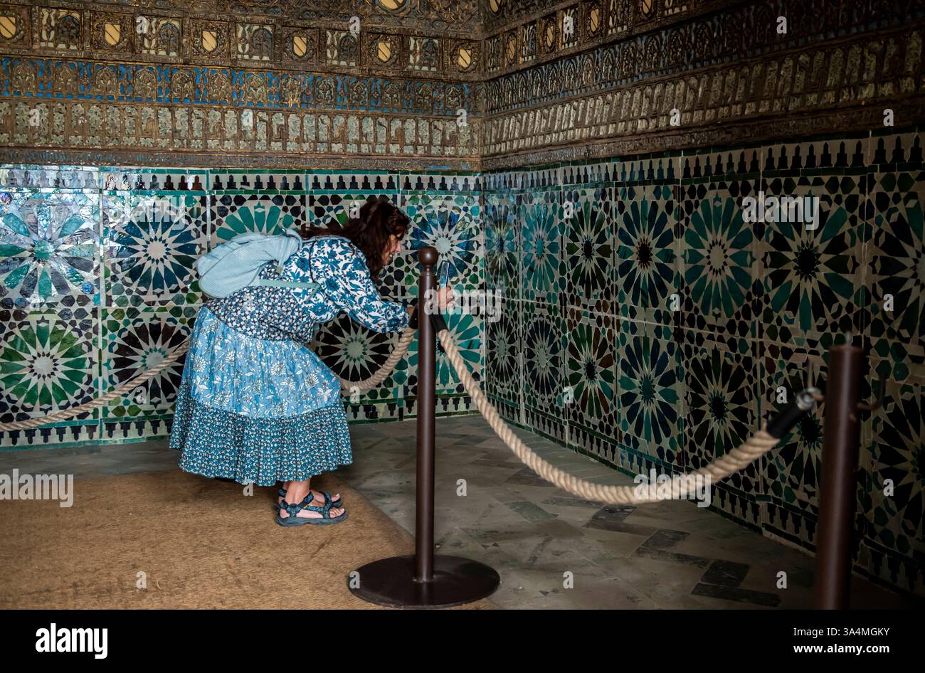 Eine Frau in einem Patchworkkleid mit grünen und blauen Blumenmustern beobachtet und fotografiert wunderschöne alte Fliesen in einer Kapelle in Corboba in Spanien. Die Muster und Farben ihres Kleides passen unheimlich zu den Fliesen, die sie ansieht. Stockfoto