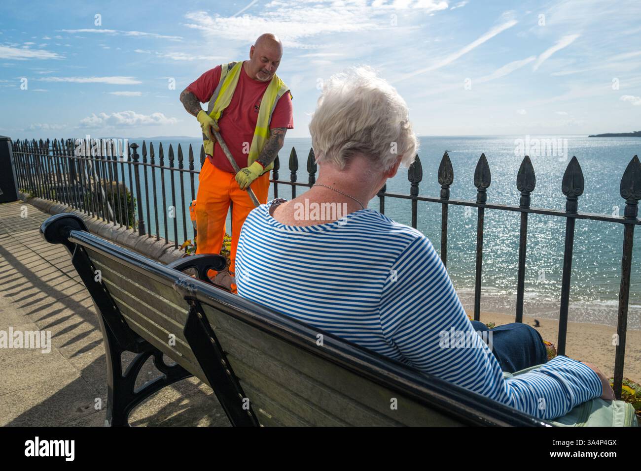 Tenby, Südwales, Großbritannien: Ein Arbeiter, der als ältere Dame auffegt, sitzt auf einer Bank mit Blick auf das Meer. Stockfoto