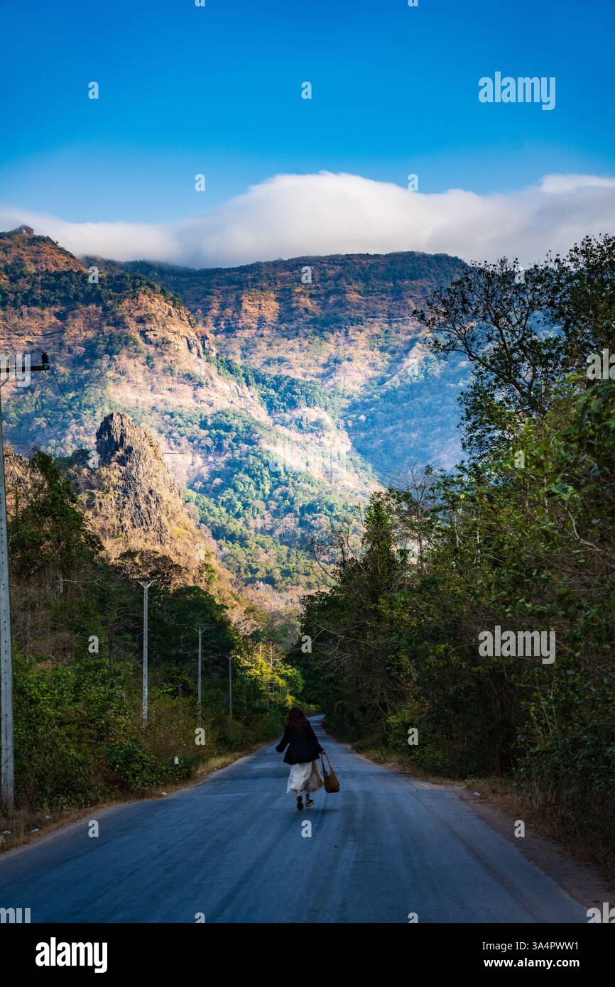 Frau, die auf einer malerischen Bergstraße in Südostasien spaziert, umgeben von üppigem Grün und dramatischen Gipfeln unter einem hellblauen Himmel Stockfoto