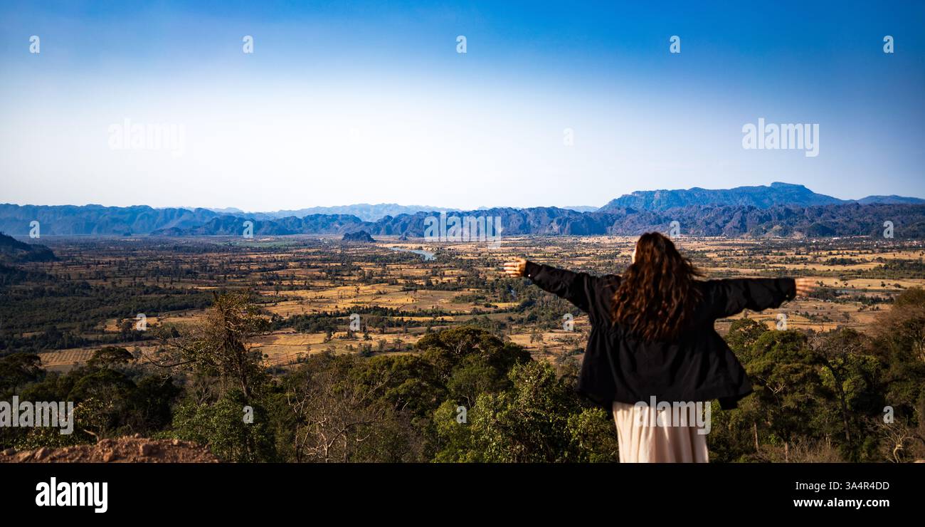 Frau, die auf einem Aussichtspunkt in den Bergen steht, Arme weit offen und das weite Tal und die zerklüfteten Gipfel in Südostasien bewundert. Stockfoto
