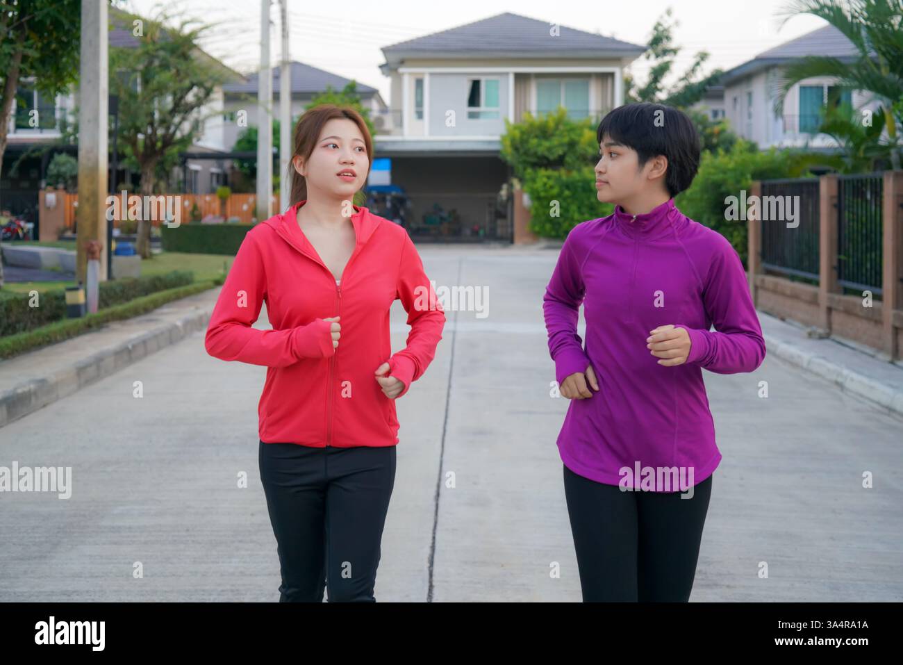 Zwei asiatische Freunde joggen Seite an Seite in einem ruhigen Vorstadtbereich, gekleidet in hellen Laufjacken und Aktivkleidung. Stockfoto