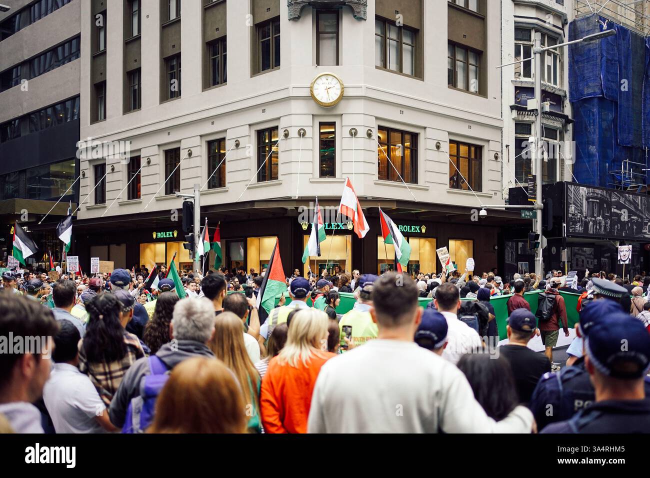 Ein dynamischer Protest im CBD von Sydney: Palästinensische Fahnen wehen inmitten der verschwommenen Menschenmassen in der Nähe des Rolex-Geschäfts, eingerahmt von einem historischen Uhrenturm und Zuschauern. Stockfoto
